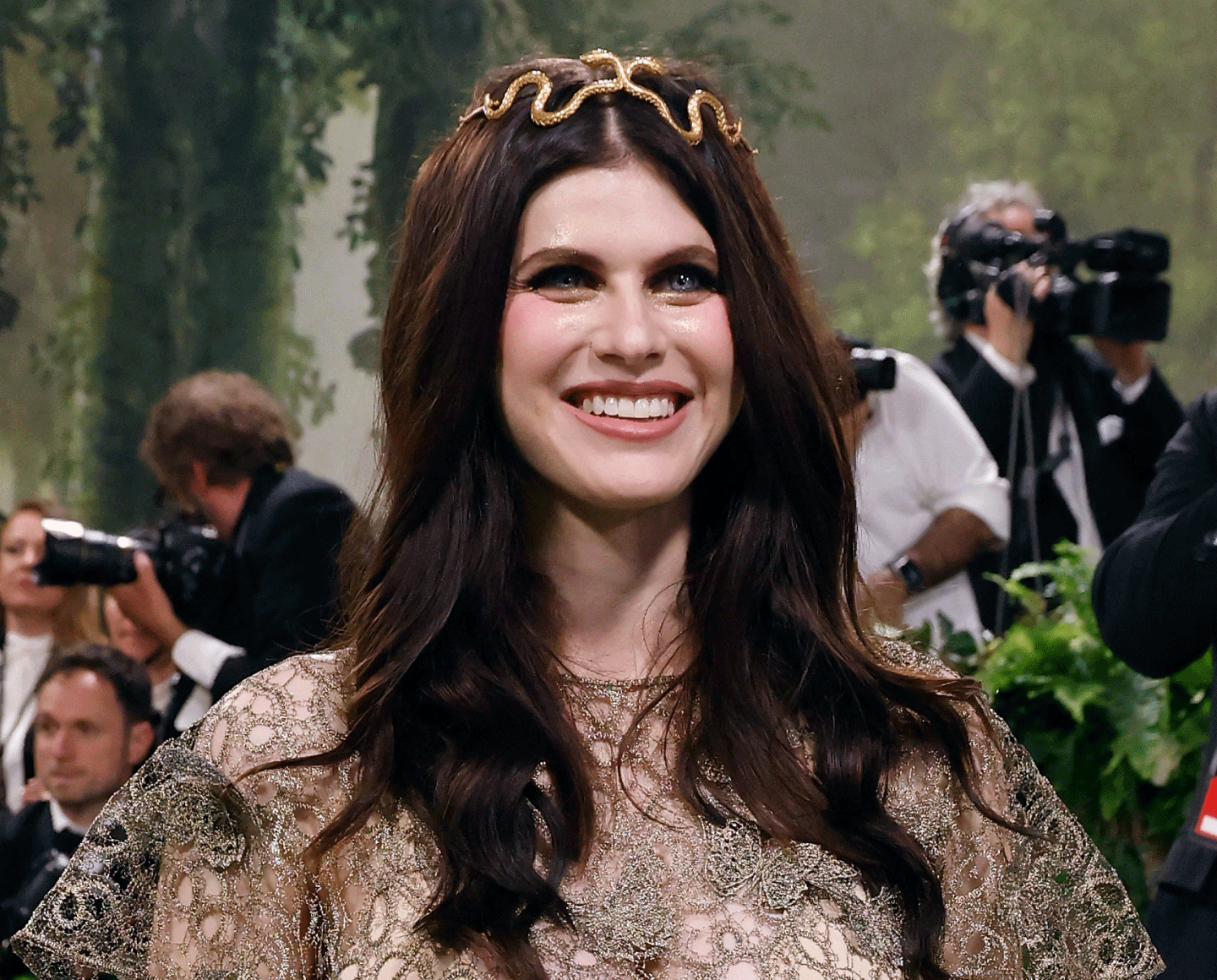 A woman with long brown hair wearing a gold snake-shaped headband and a sheer, ornate dress smiles while surrounded by photographers and lush greenery at a formal event.