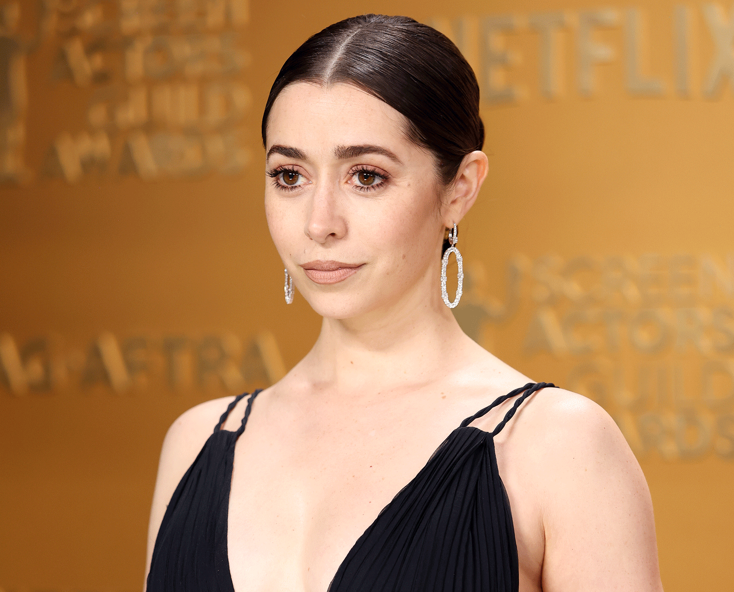 A woman with dark hair tied back and wearing drop earrings and a black dress stands in front of a gold backdrop with blurred text.