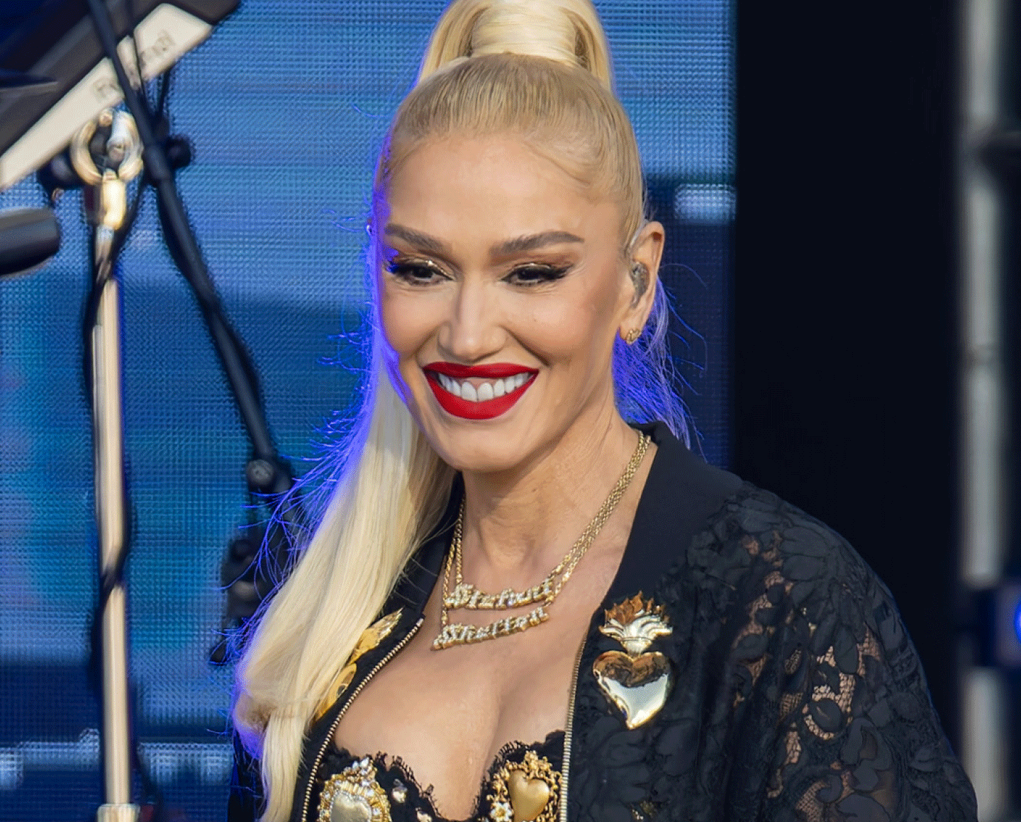 A blonde woman with long hair in a high ponytail, wearing a black lace outfit with gold details and a bold red lipstick, smiles on stage with a microphone and blue lights in the background.