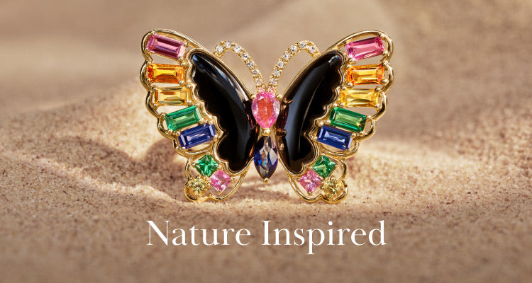 A jeweled butterfly brooch with colorful gemstones and black wings sits on sand. The words Nature Inspired are written below the brooch.