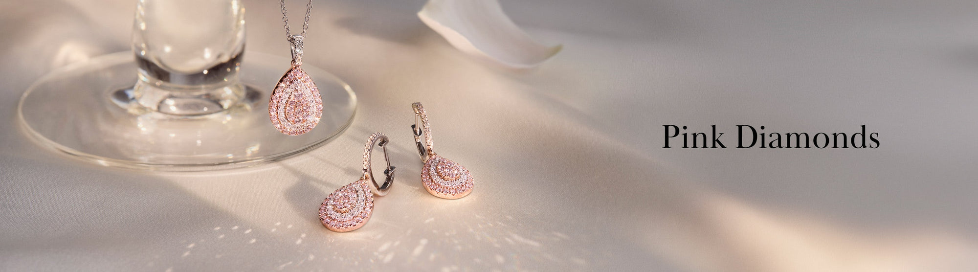 Elegant pink diamond pendant and earrings set displayed on a soft surface beside a glass, with the words Pink Diamonds written on the right side.