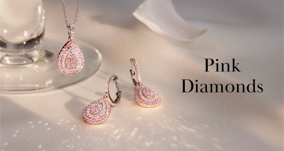 A necklace and matching earrings, each with teardrop-shaped pink diamond settings, are displayed on a light surface next to a glass and a white flower petal. The text reads: Pink Diamonds.