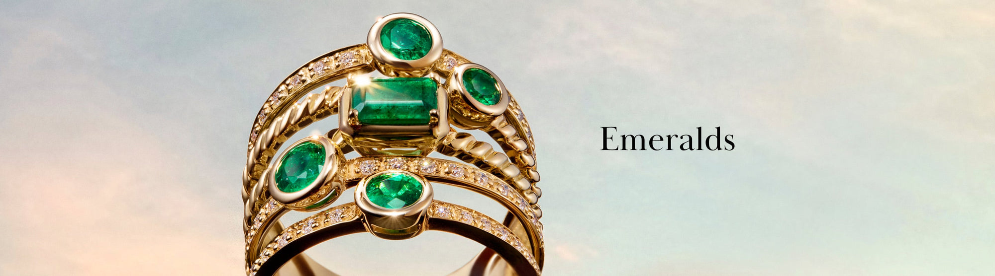Five gold rings with green emerald gemstones and small diamonds stacked together, set against a soft sky background, with the word Emeralds written beside them.