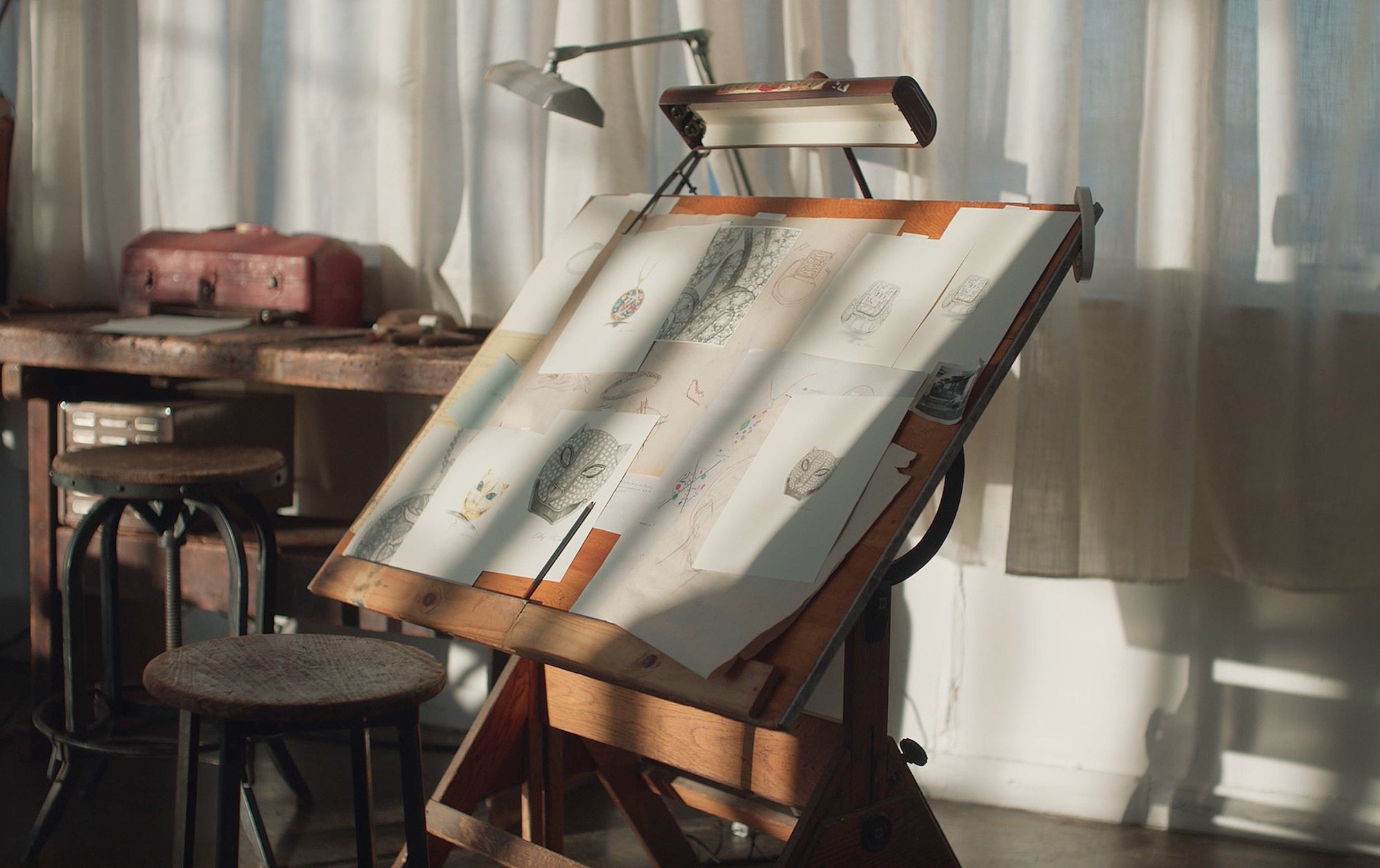 A wooden drafting table with sketches and drawings on paper, lit by sunlight from a nearby window. A lamp, wooden stools, a suitcase, and other art supplies are in the background, creating a cozy, artistic workspace.