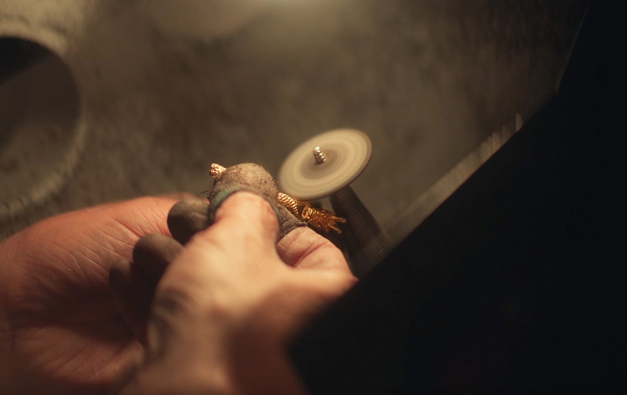 Close-up of hands holding and polishing a small metal object with a rotary tool, with motion blur on the spinning wheel. The lighting is warm and focused on the workspace.