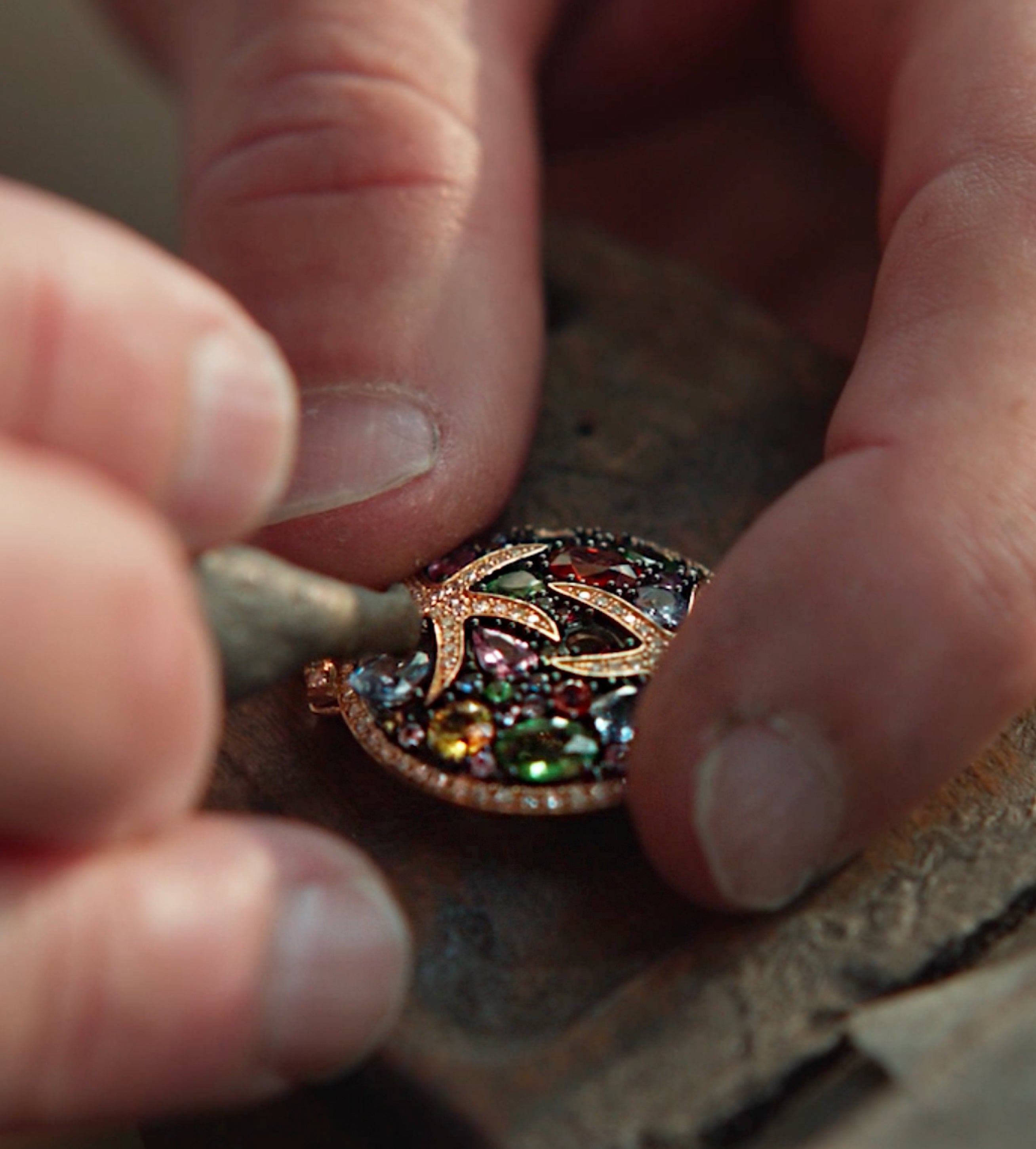 A close-up of hands working on an ornate, colorful pendant with a bird design, using a small tool for detailed craftsmanship.