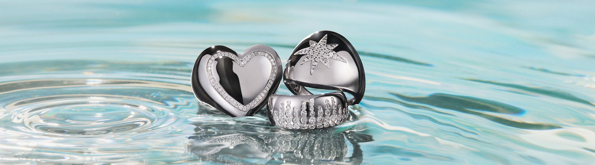 Three shiny silver rings, including a heart, a starburst design, and a textured band, rest on rippling water with a soft blue background. The word Silver appears on the right.