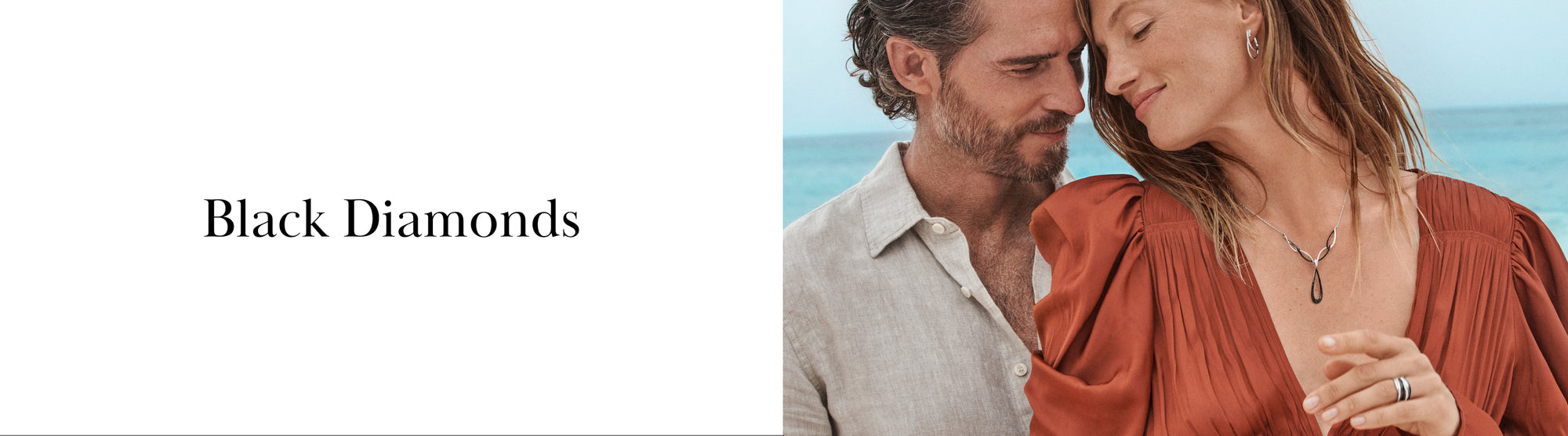 A couple stands close together by the sea; the woman wears a flowing rust-colored dress and a necklace with black diamond jewelry. The text Black Diamonds appears on a white background.