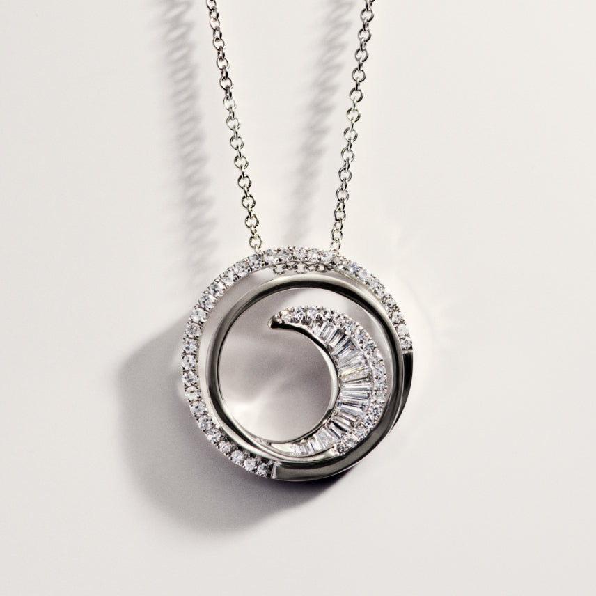 A silver necklace with a circular pendant featuring a crescent shape encrusted with diamonds inside a diamond-studded outer ring, displayed on a light background.