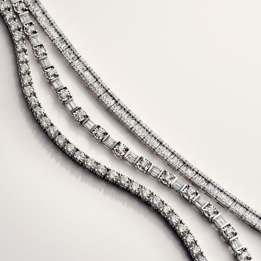 Four elegant diamond tennis bracelets are arranged diagonally on a light surface, showcasing different styles of diamond cuts and settings, with sparkling stones catching the light.