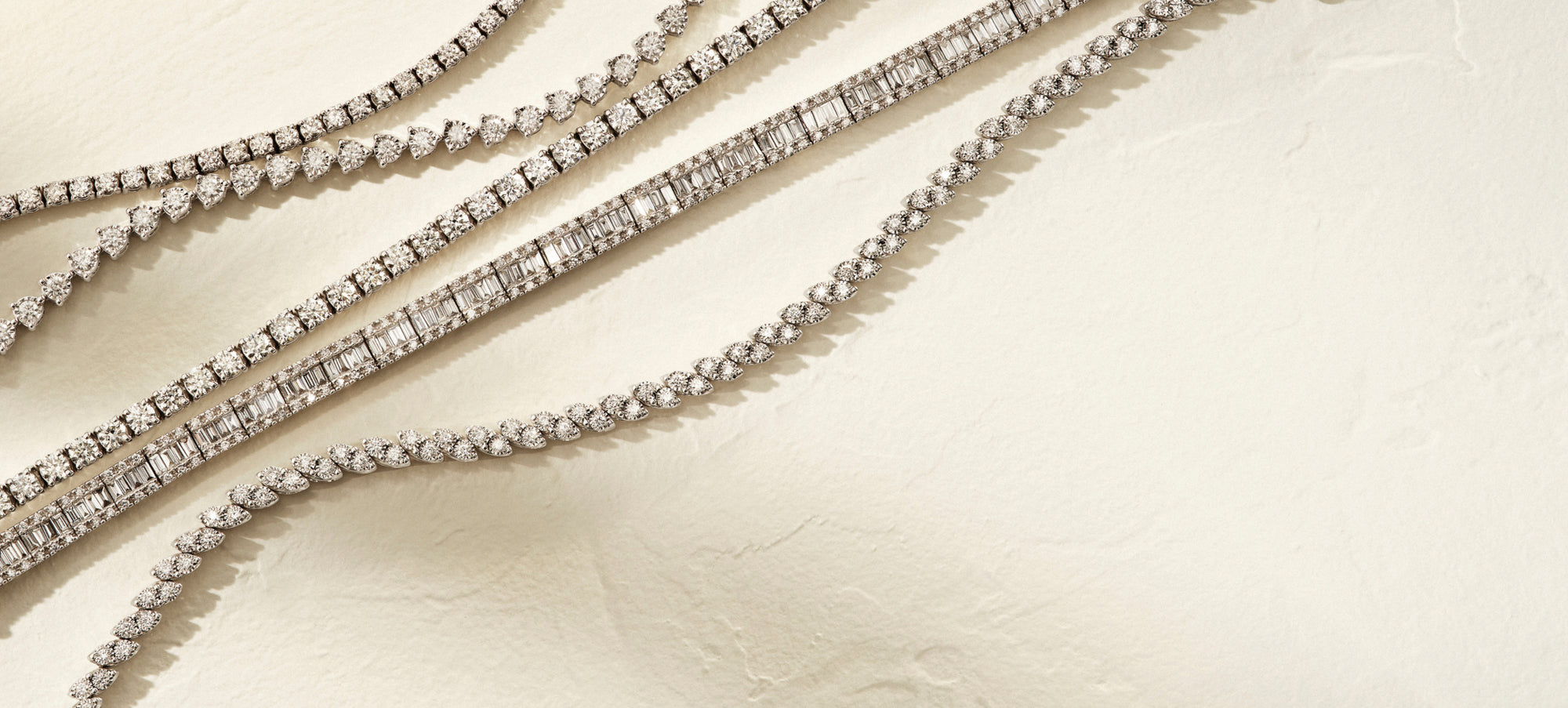 Four elegant diamond necklaces are arranged diagonally on a textured, light beige surface. Each necklace features a different design, with sparkling diamonds set in various patterns.