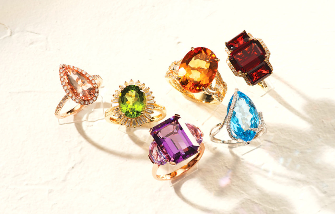 Six gemstone rings with various colorful stones—pink, green, orange, red, blue, and purple—are arranged on a light textured surface, casting soft shadows.