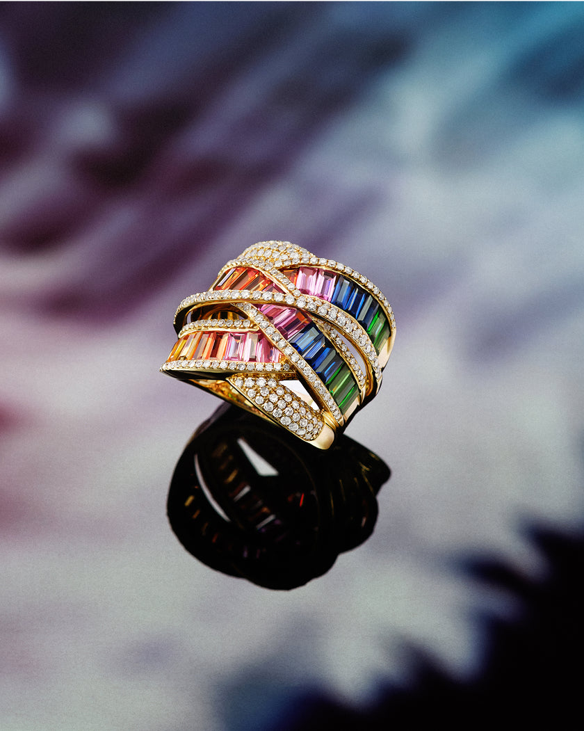 A gold ring featuring colorful, rectangular gemstones arranged in wavy bands, accented with rows of small white diamonds, is displayed against a blurred, multicolored background with its reflection visible below.