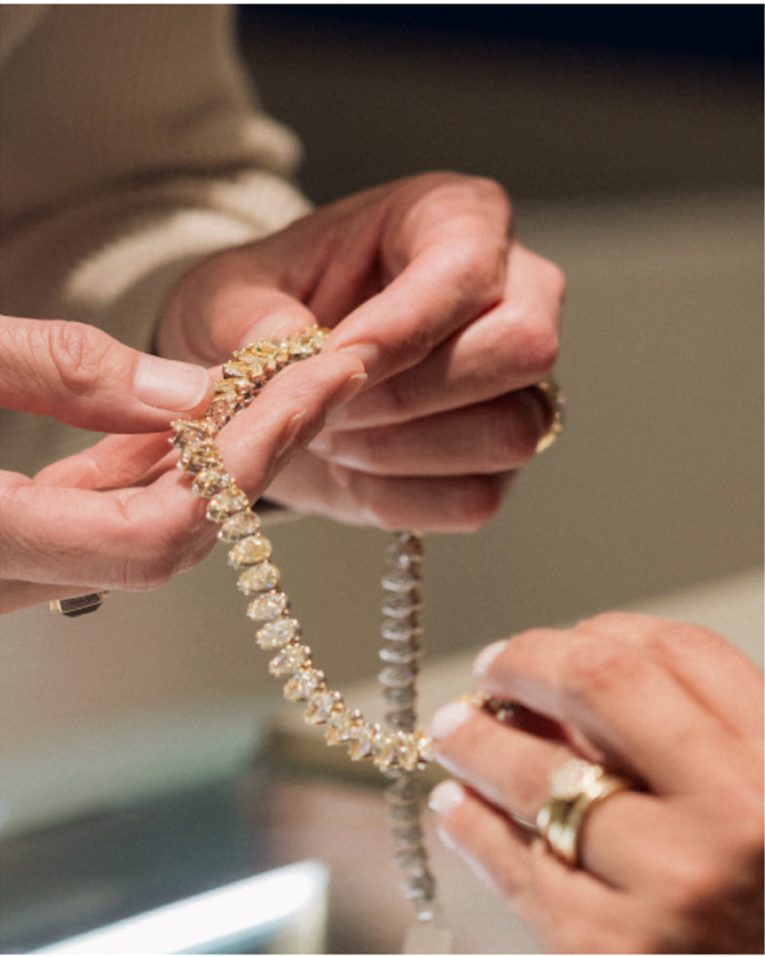 Two people examine a diamond bracelet, each holding one end. The hands are adorned with gold rings and the image is softly lit, highlighting the sparkling jewelry.