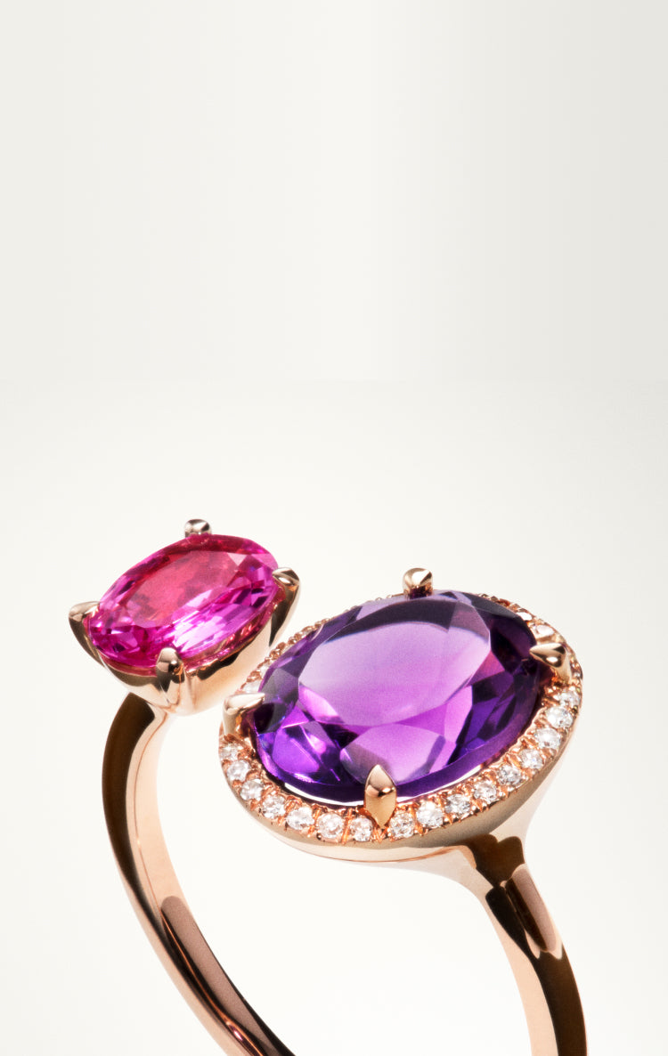A rose gold ring featuring two stones: one oval purple gemstone surrounded by small diamonds and one smaller oval pink gemstone, both set in an open band design against a light background.