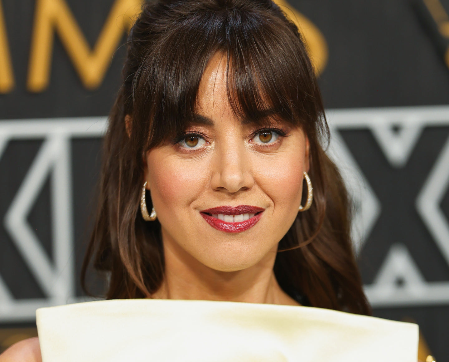 A woman with medium-length brown hair and bangs smiles at the camera. She wears hoop earrings, glossy lipstick, and a cream-colored outfit, with a blurred black and gold background behind her.