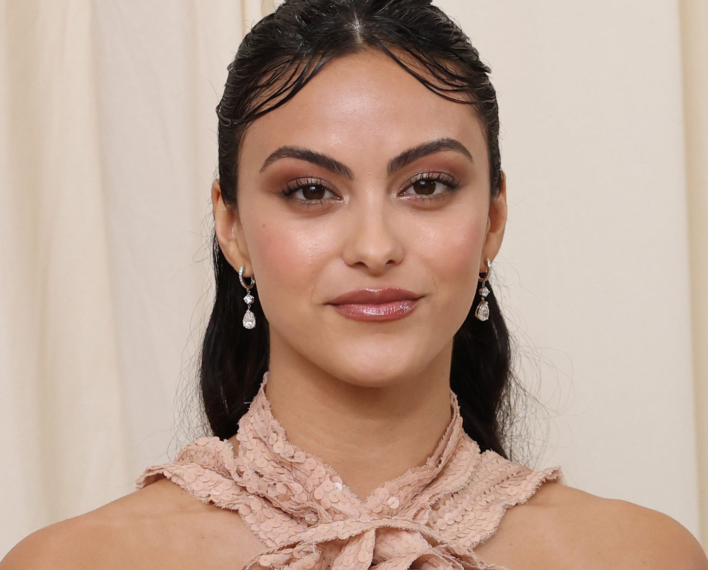 A woman with dark, wavy hair styled back and subtle curls at the front wears drop earrings and a textured, light pink halter top. She has natural makeup and a slight smile, standing against a cream-colored background.