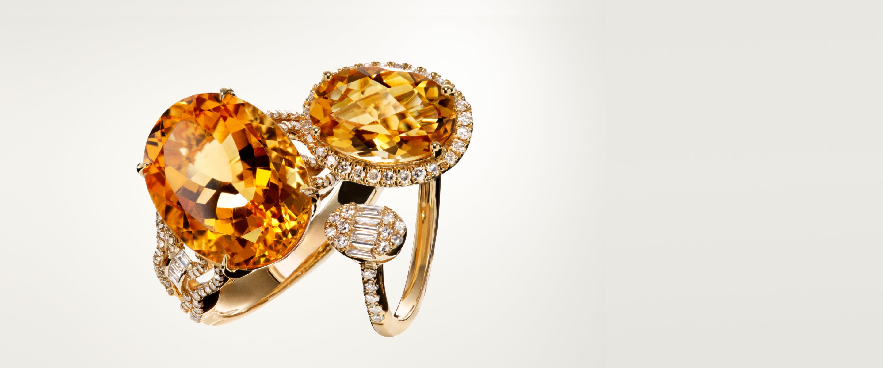 Two gold rings, each featuring a large oval orange gemstone surrounded by small white diamonds, with one ring also accented by a round, diamond-encrusted detail on the band, displayed on a light background.