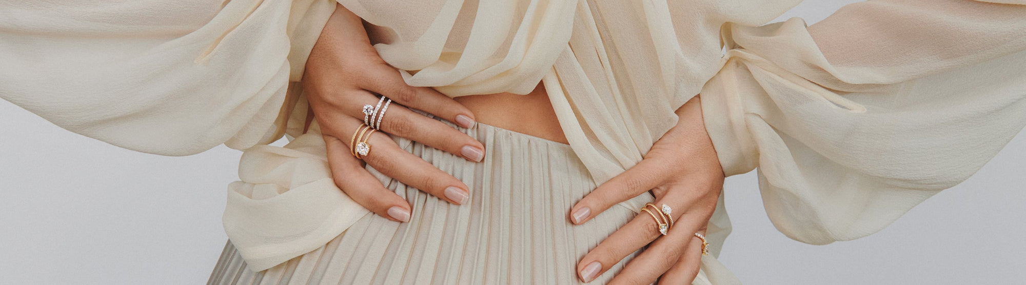 A person wearing a sheer, flowy cream blouse and pleated skirt, with their hands on their waist, showcasing several elegant rings on their fingers.