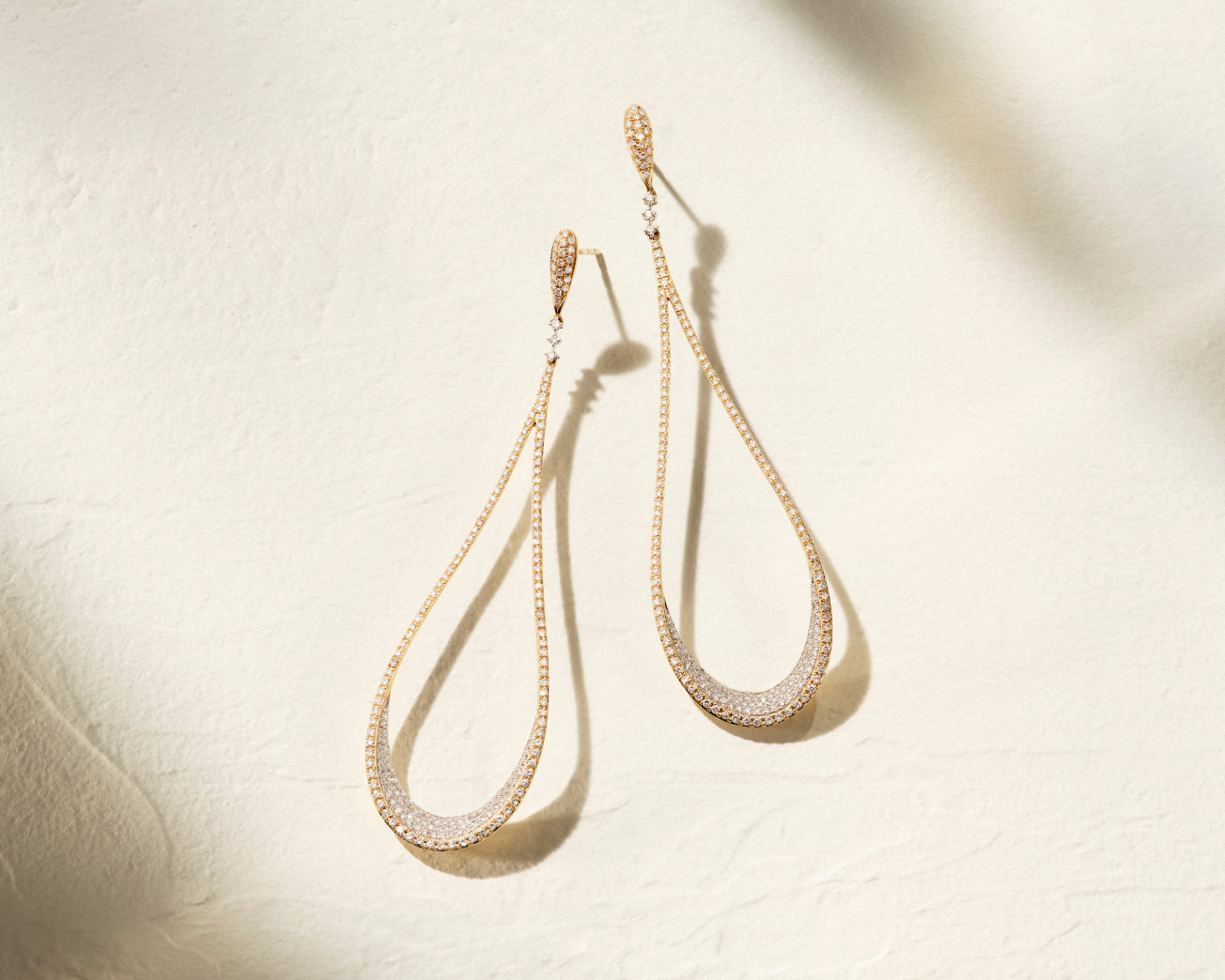 A pair of long, gold teardrop-shaped earrings with small embedded crystals, displayed against a soft beige textured background with subtle shadows.