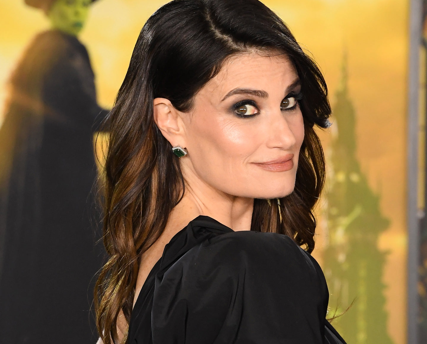 A woman with long brown hair and dramatic eye makeup looks over her shoulder and smiles slightly. She is wearing a black outfit, with a blurred green figure in the yellowish background.