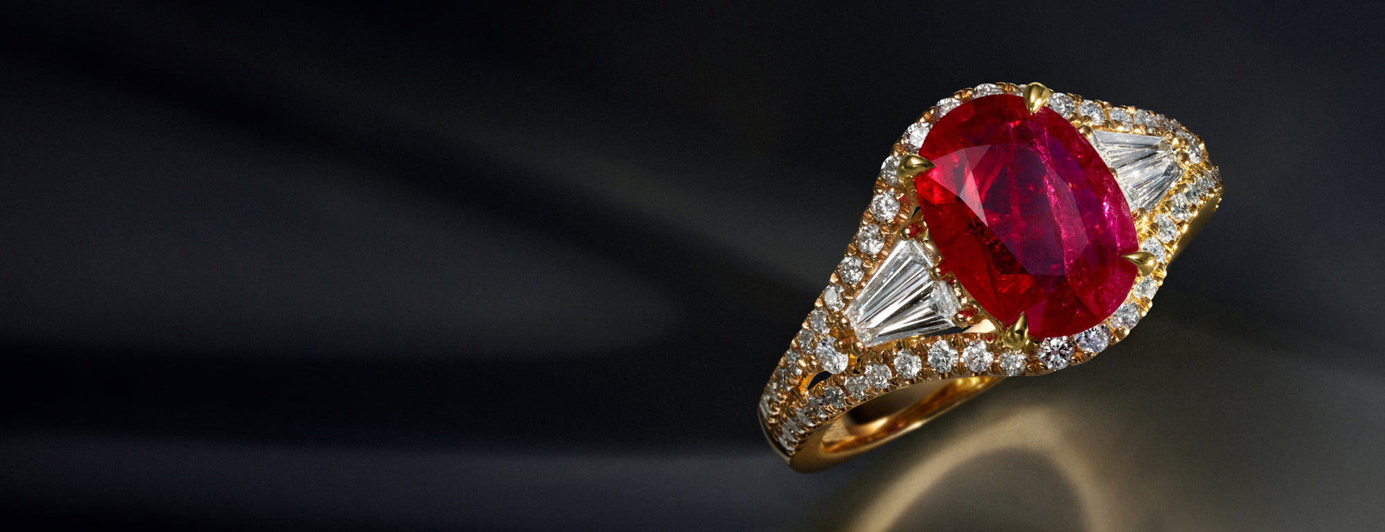 A gold ring featuring a large oval red gemstone, flanked by two triangular clear stones and surrounded by small round diamonds, displayed on a dark, reflective surface.