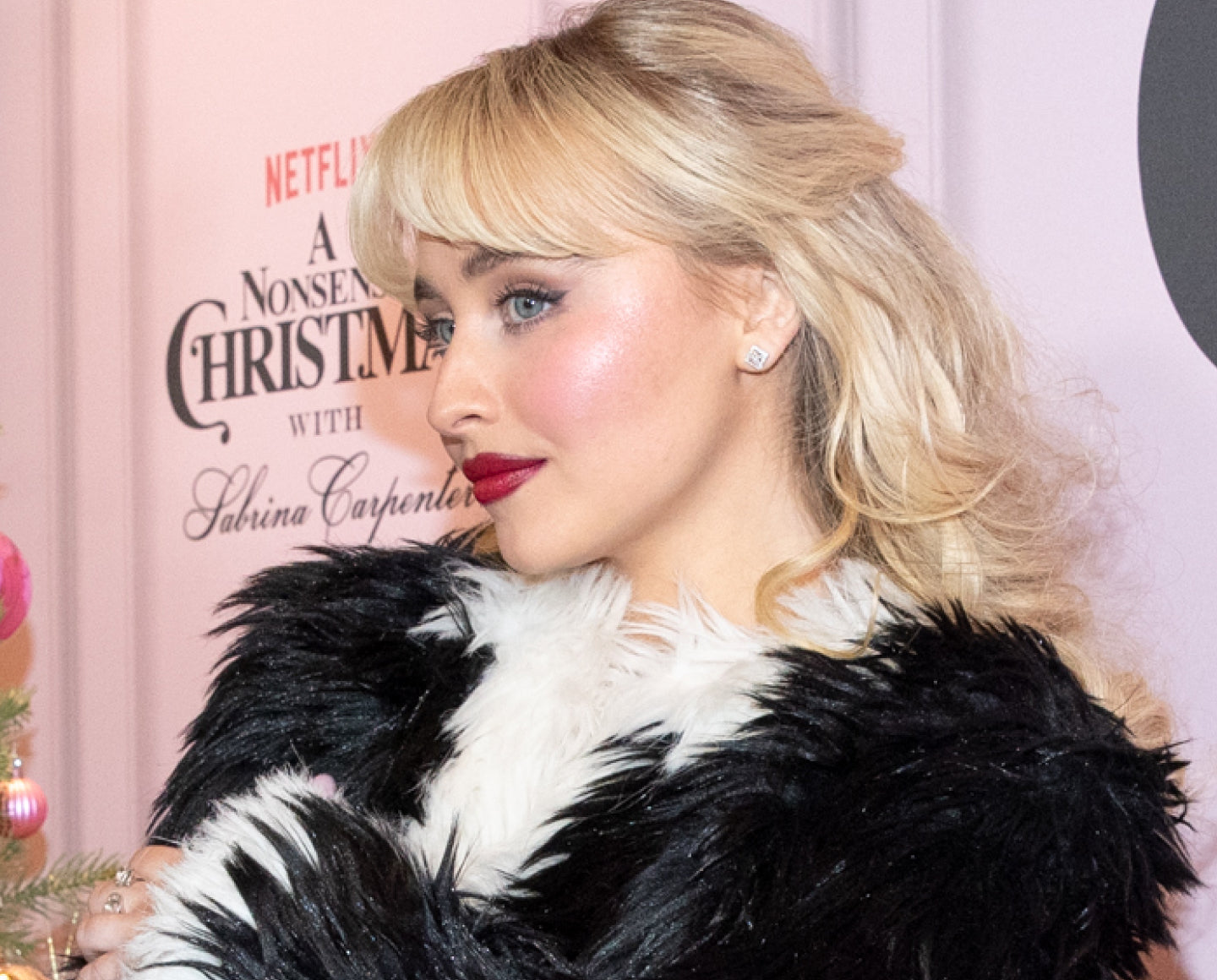 A woman with blonde hair and red lipstick poses in profile, wearing a black and white fur coat. Behind her is a sign that reads A Nonsense Christmas with Sabrina Carpenter and Netflix branding.
