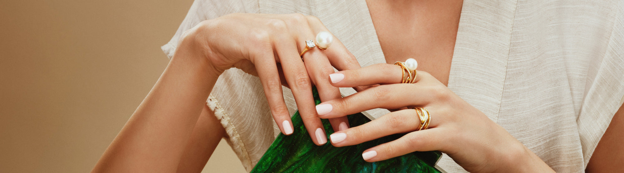 A person wearing a cream-colored blouse displays several gold rings, some with pearls or a diamond, on well-manicured hands holding a green textured object.