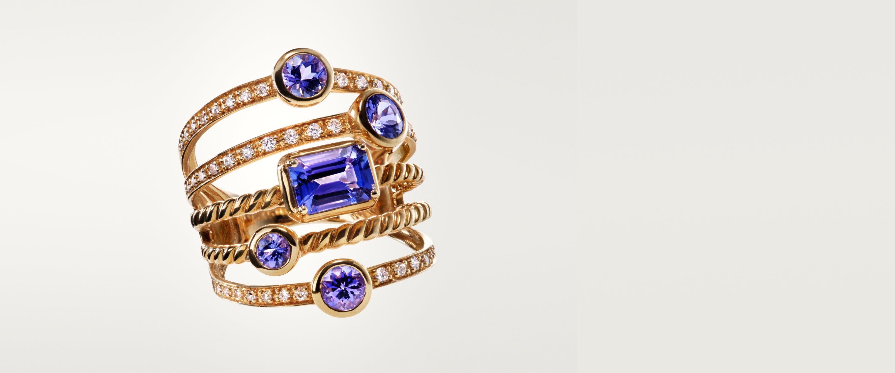 A gold ring featuring multiple intertwined bands, adorned with small diamonds and five prominent blue gemstones in various shapes and sizes, set against a plain light background.