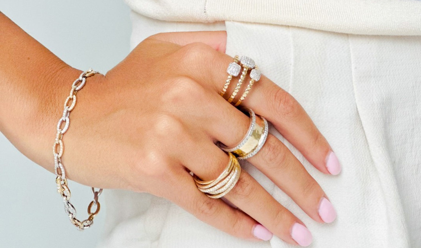 A hand with light pink nail polish wears multiple gold and silver rings with diamonds, and a matching chain bracelet, resting on cream-colored pants.