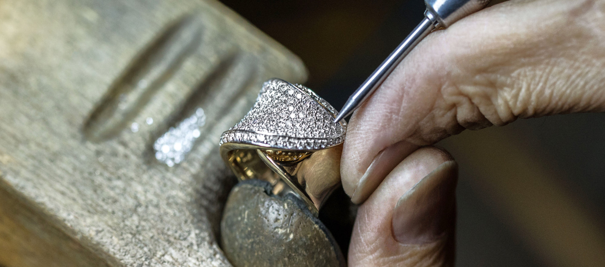 A close-up of a jeweler’s hand using a tool to set small diamonds into an ornate ring, with loose diamonds and a wooden workbench visible in the background.