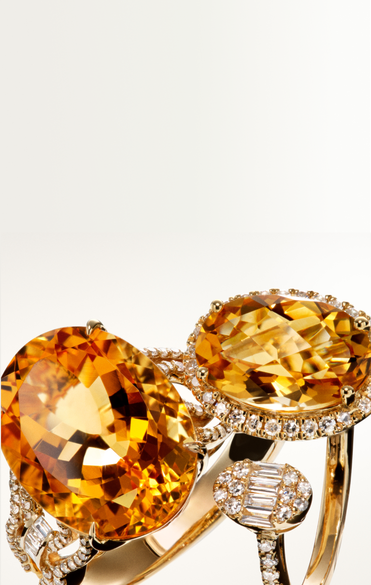 Close-up of three gold rings, each featuring large, oval-cut yellow-orange gemstones surrounded by small diamonds, displayed against a light, gradient background.