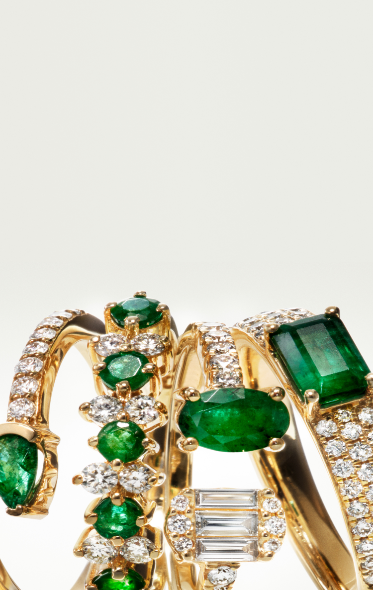 A close-up of five gold rings adorned with green emeralds and sparkling diamonds, arranged in a row against a plain light background.
