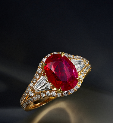A gold ring featuring a large oval red gemstone at the center, flanked by baguette-cut clear stones and surrounded by small round diamonds, all set against a dark background.